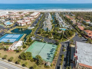 An aerial view of a coastal resort with multiple buildings, a large swimming pool, tennis courts, and a beach in the background. - Sea Winds 51 by Distinctive Beach Rentals (St. Augustine)