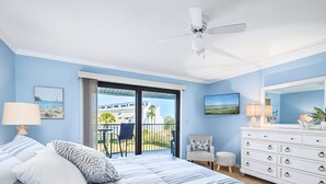 A bedroom with a bed, a dresser, a chair, a lamp, and a balcony with a view of buildings and palm trees. - Sea Winds 51 by Distinctive Beach Rentals (St. Augustine)