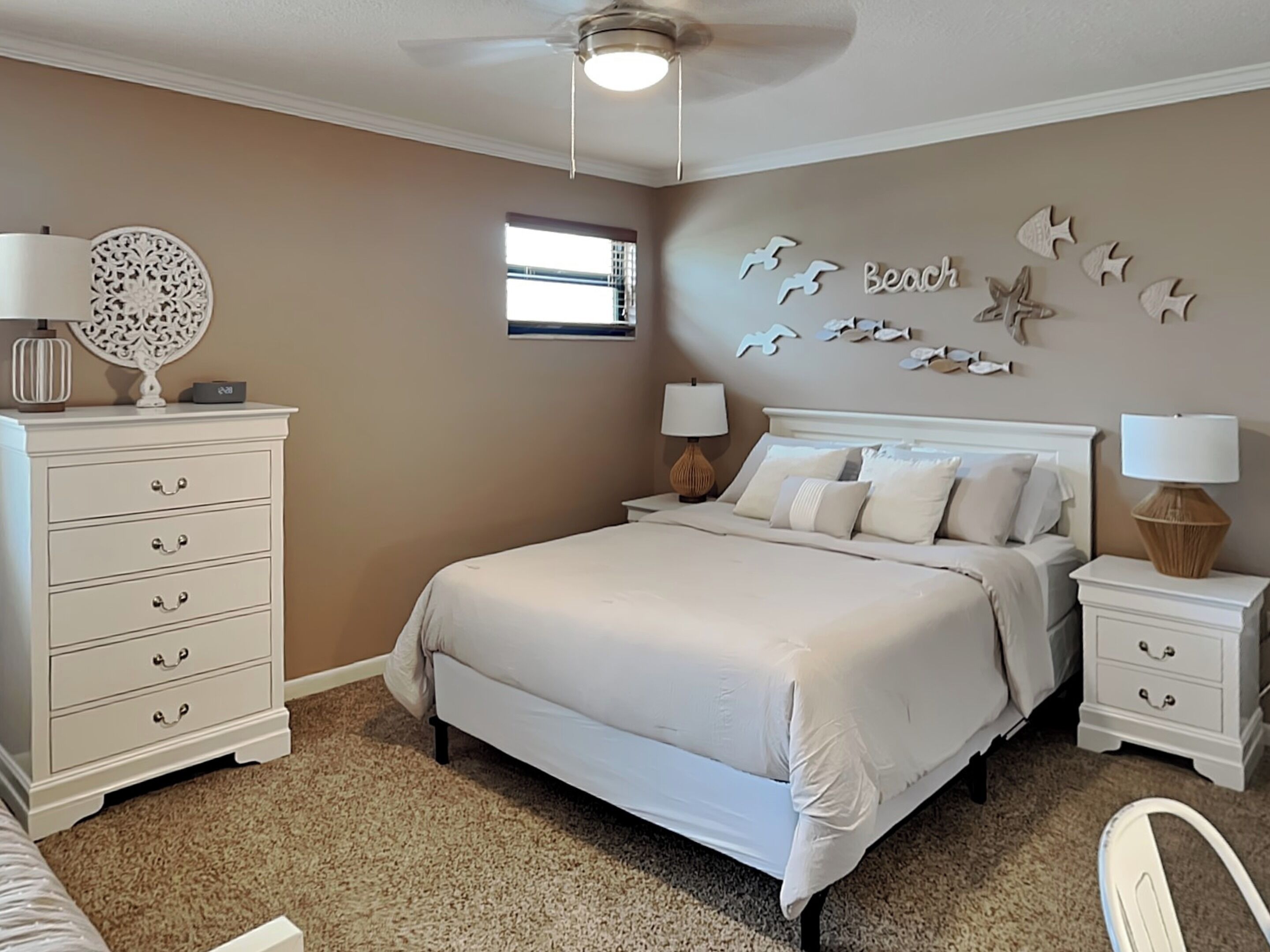 A bedroom with a large bed, two bedside tables with lamps, a dresser, a ceiling fan, and a wall decoration with seagulls and fish.