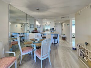 Een moderne keuken met een centraal eiland, witte kasten en een eethoek met tafels en stoelen met glazen blad.