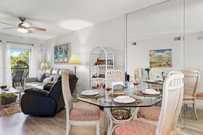 Een eetruimte met een glazen tafel, witte stoelen en een plafondventilator.