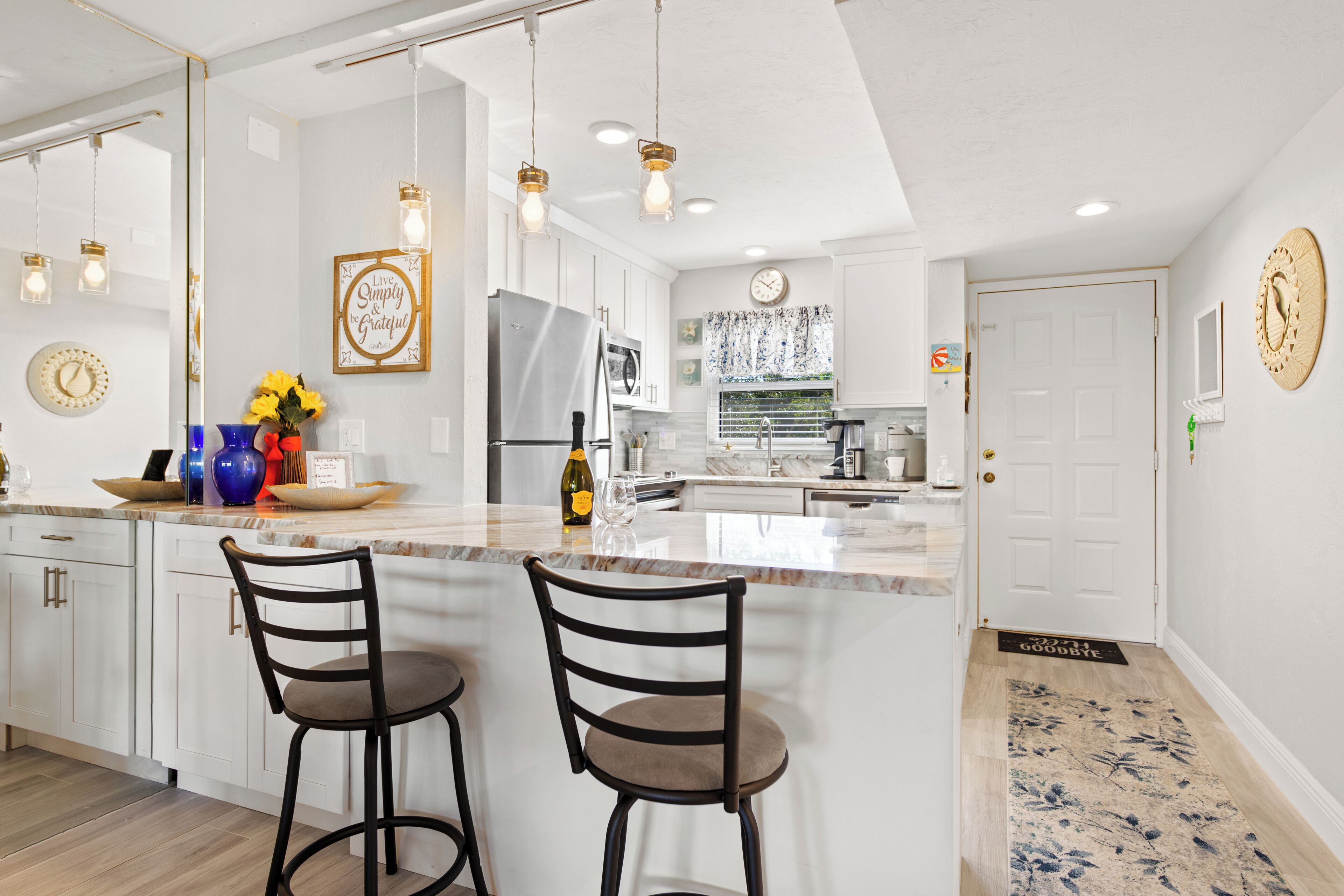 A modern kitchen with white cabinetry, a stainless steel refrigerator, and a marble countertop. There are two bar stools and a decorative wall clock.