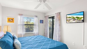 Een slaapkamer met een bed, een plafondventilator, een raam met gordijnen en een televisie aan de muur.