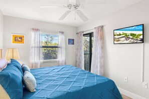 Een slaapkamer met een bed, een plafondventilator, een raam met gordijnen en een televisie aan de muur.