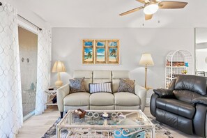 A living room with a beige sofa, a black leather chair, a glass coffee table, and a ceiling fan. There are three framed pictures on the wall and a lamp on a side table.