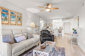 A living room with a beige sofa, a black leather chair, a glass coffee table, and a ceiling fan.