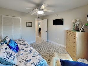 A bedroom with a bed, a dresser, a ceiling fan, a TV, and a door leading to another room.