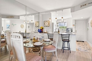 Uma área de refeições moderna com mesa de vidro, cadeiras brancas e cozinha ao fundo.