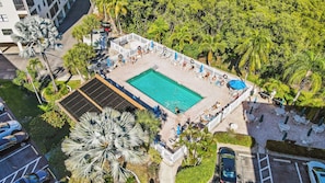 Vista aérea de um resort com piscina central cercada por cadeiras de praia e guarda-sóis, palmeiras e um estacionamento.