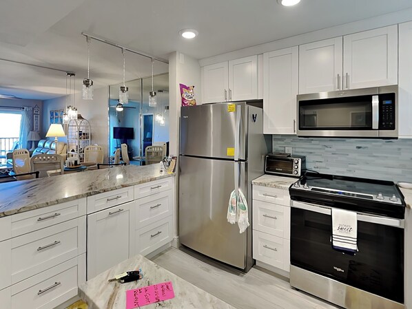A modern kitchen with white cabinets, stainless steel appliances, a marble countertop, and a large window.