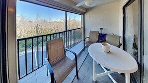 Een balkon met een witte tafel en twee stoelen, een plafondventilator en uitzicht op een weg en bomen.