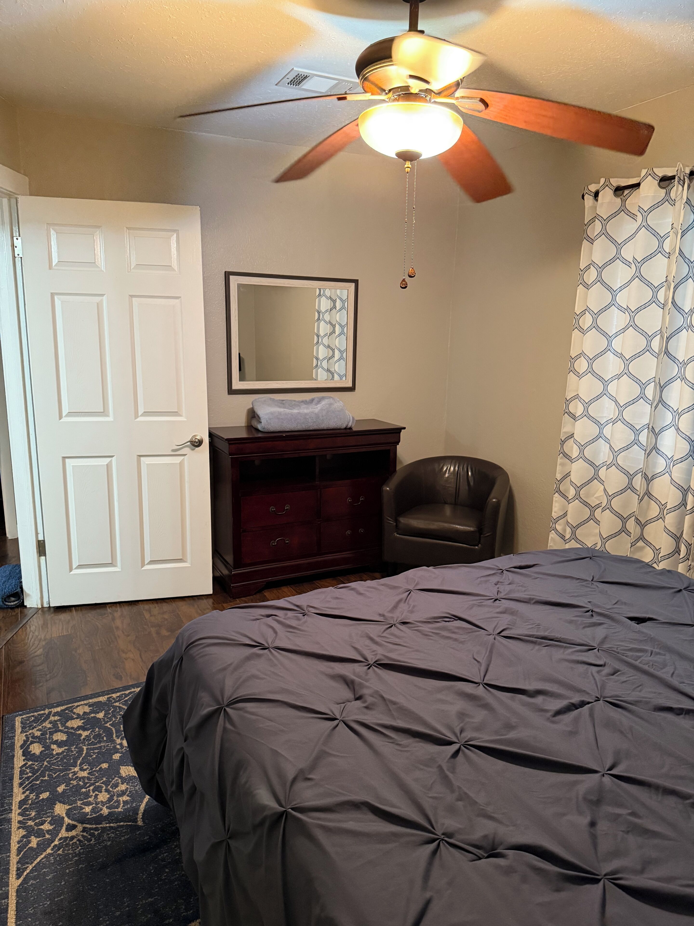 A bedroom with a bed, a dresser, a chair, a ceiling fan, and a mirror.