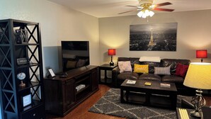 A living room with a TV, dark wood furniture, a ceiling fan, and a cityscape painting on the wall.