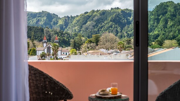 Un balcone con sedie in vimini e un tavolino, con vista su un borgo con una chiesa e dolci colline verdi.