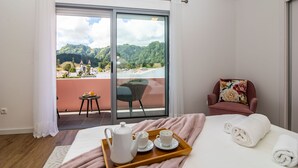 Una camera d'albergo con un letto, un balcone affacciato su un paesaggio montuoso, un tavolino con un vassoio di prodotti per la colazione e una sedia con un cuscino decorato con fiori.