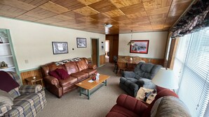 A cozy living room with a wooden ceiling, a brown leather couch, a plaid armchair, a red armchair, a wooden coffee table, and a dining table with chairs.