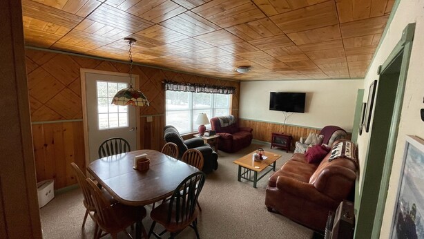 A cozy cabin interior with a wooden ceiling, a dining table with chairs, a sofa, and a television mounted on the wall.