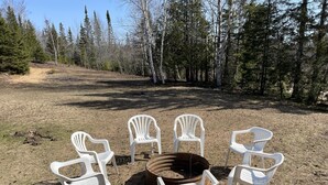 A group of white plastic chairs arranged around a central fire pit in an outdoor setting with a dirt ground and trees in the background.