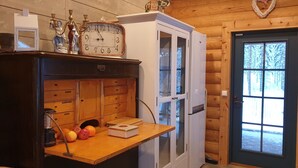 Interieur van een houten hutje met een houten tafel, een klok, een koelkast en een deur naar buiten.