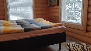 Een gezellig houten blokhut slaapkamer met een bed, een raam met luxaflex en uitzicht op besneeuwde bomen.