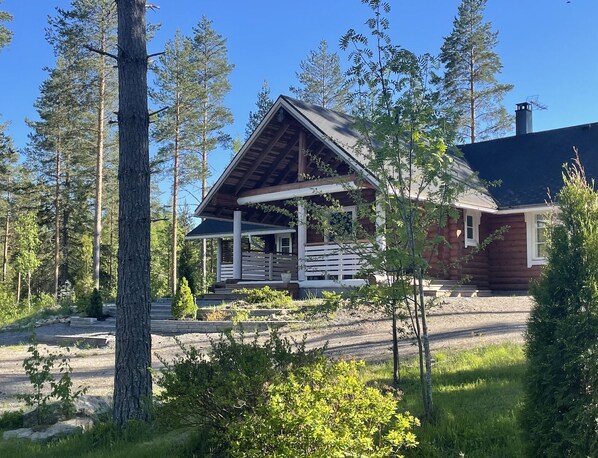 Hirsimökki, jossa on kuisti, ja sen ympärillä on puita sekä sorapäällysteinen ajotie.