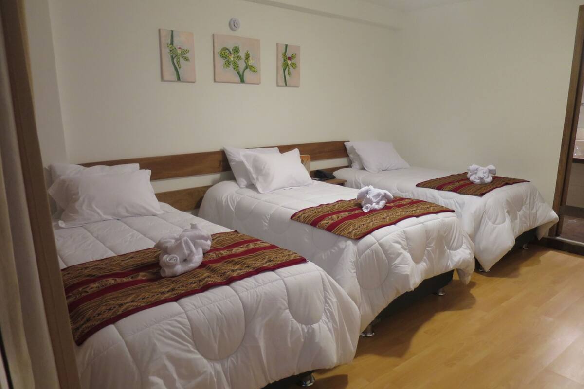 Une chambre d’hôtel avec trois lits simples, chacun doté de draps blancs et d’une couverture à motifs rouges et dorés. Trois tableaux représentant des fleurs sont accrochés au mur au-dessus des lits.
