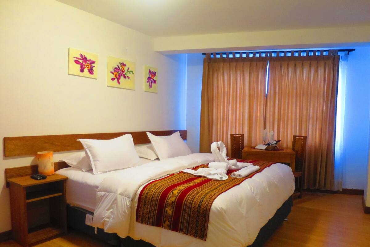 Une chambre d’hôtel avec un grand lit, une table de chevet avec une lampe, une tête de lit en bois et trois tableaux aux motifs floraux accrochés au mur.