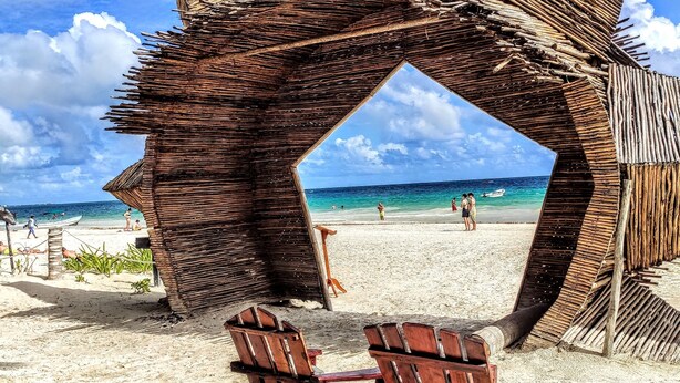 A beach with a thatched structure and two wooden chairs.