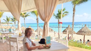 A covered outdoor seating area with white tables and chairs, overlooking a beach with thatched umbrellas and palm trees.
