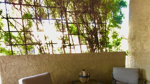 A patio area with two chairs and a small table, a textured wall, and a view of greenery.