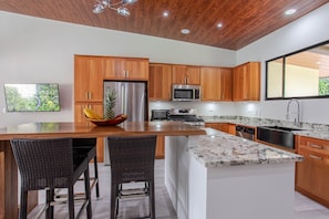 A modern kitchen with wooden cabinets, stainless steel appliances, a large island with bar stools, and a view of greenery outside. - Secluded Luxury Rainforest Villa on the Baru River close to Dominical Beach ! (Dominical)