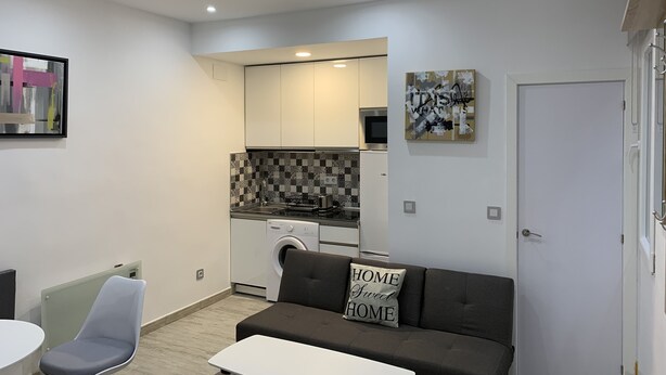 A compact living space with a kitchenette, a sofa with a 'HOME Sweet HOME' pillow, a dining area with a chair, and a white door. - Charming apartment in the old center of Madrid (Madrid)