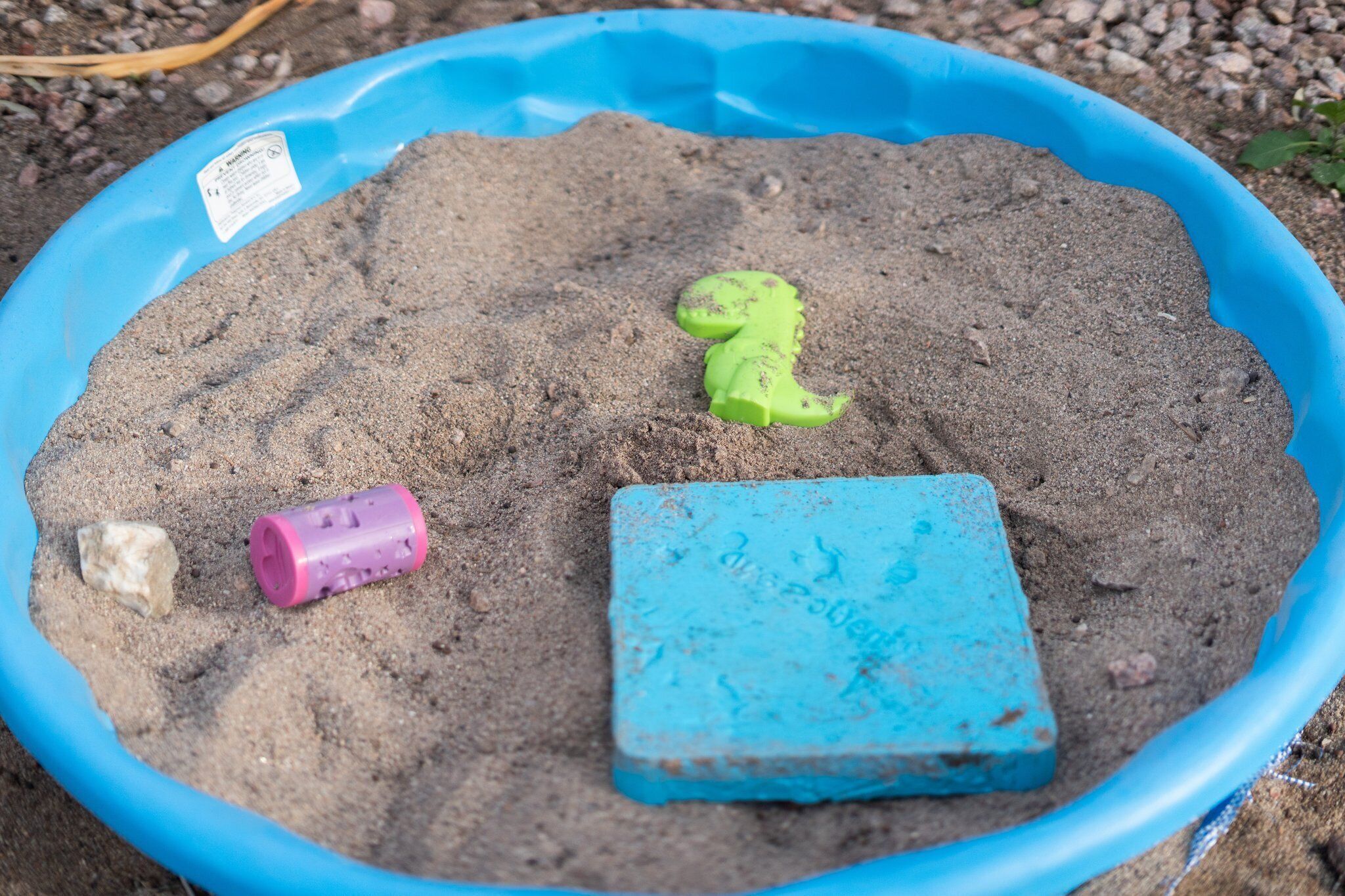 A blue plastic kiddie pool filled with sand, a green toy dinosaur, a pink toy, and a blue toy.