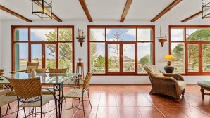 Una habitación amplia con ventanas grandes, una mesa de comedor con sillas y un sofá de mimbre.