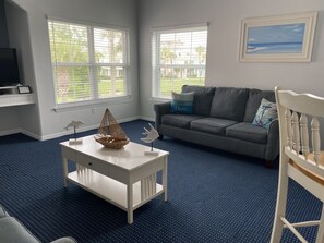 A living room with a grey sofa, a white coffee table with decorative items, and large windows with blinds.