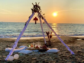 A beach setup with a canopy, a table with food and drinks, and a view of the ocean at sunset. - Marbella Beach Hotel (Golem)