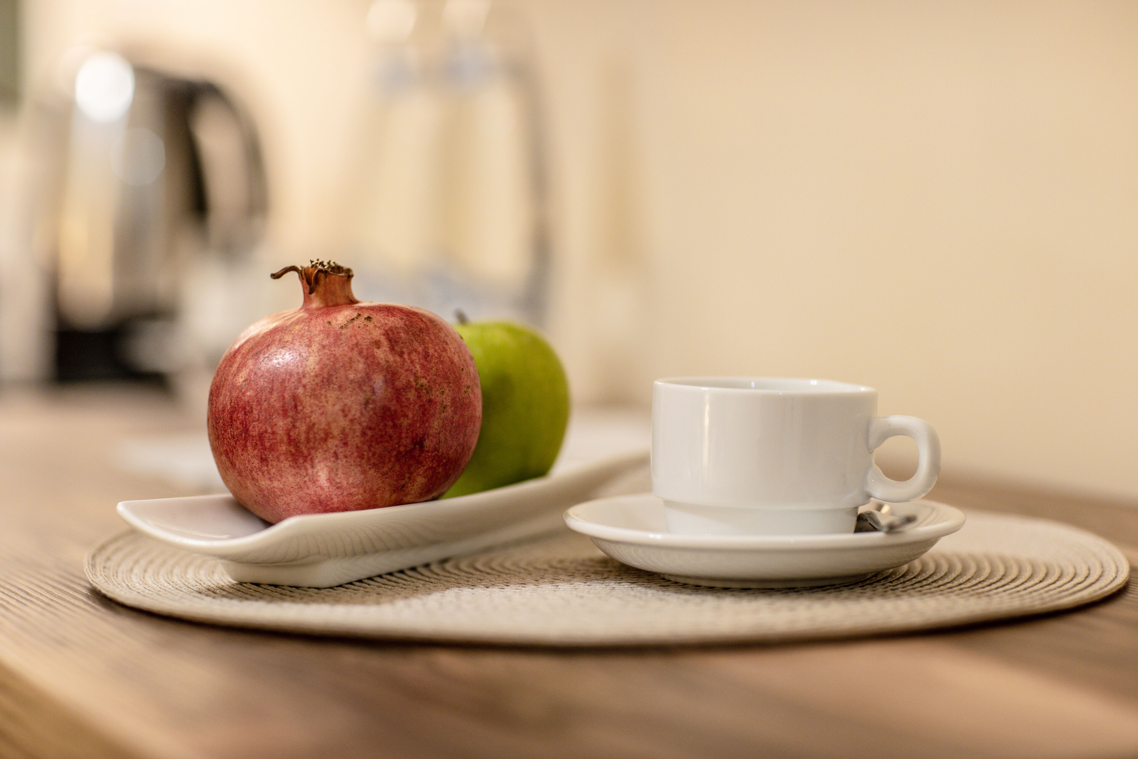 A pomegranate, a green apple, and a white cup on a table with a placemat.
