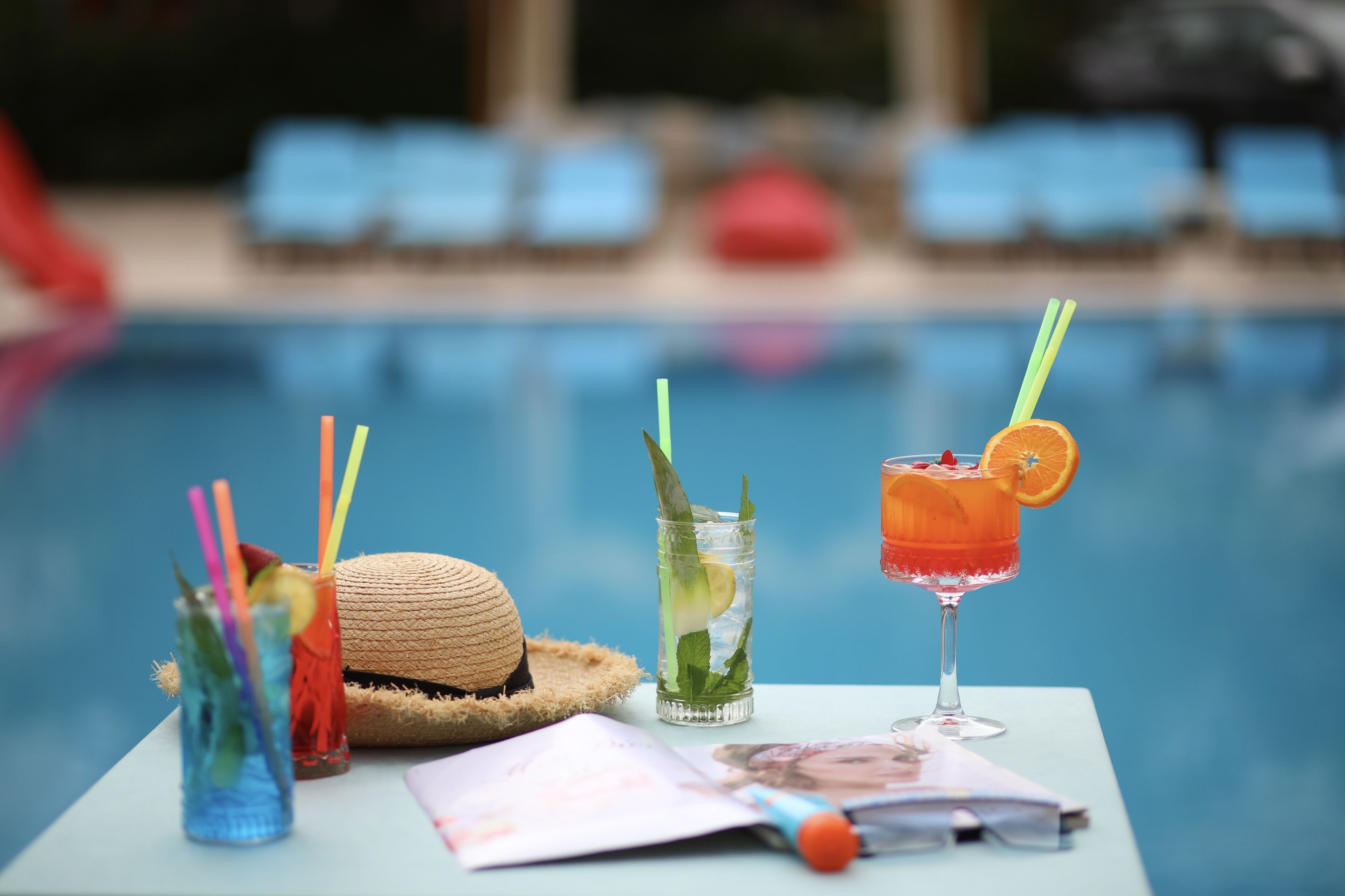 A table with drinks, a straw hat, and a magazine by a pool.