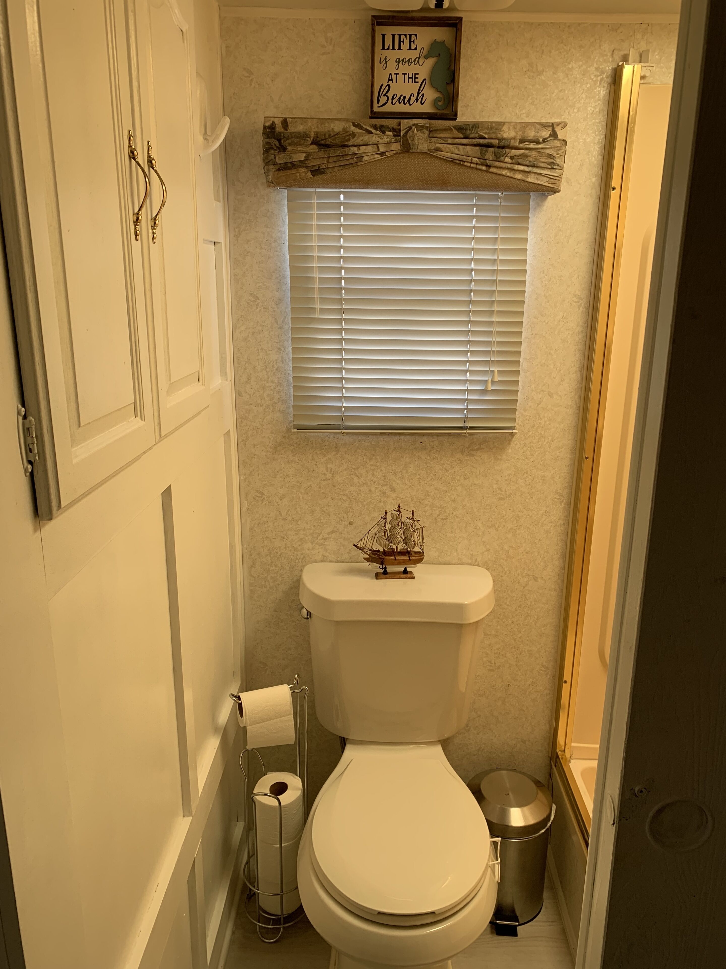 Un baño con inodoro, un rollo de papel higiénico, un adorno con forma de barco en el inodoro, una ventana con persianas y una zona de ducha.