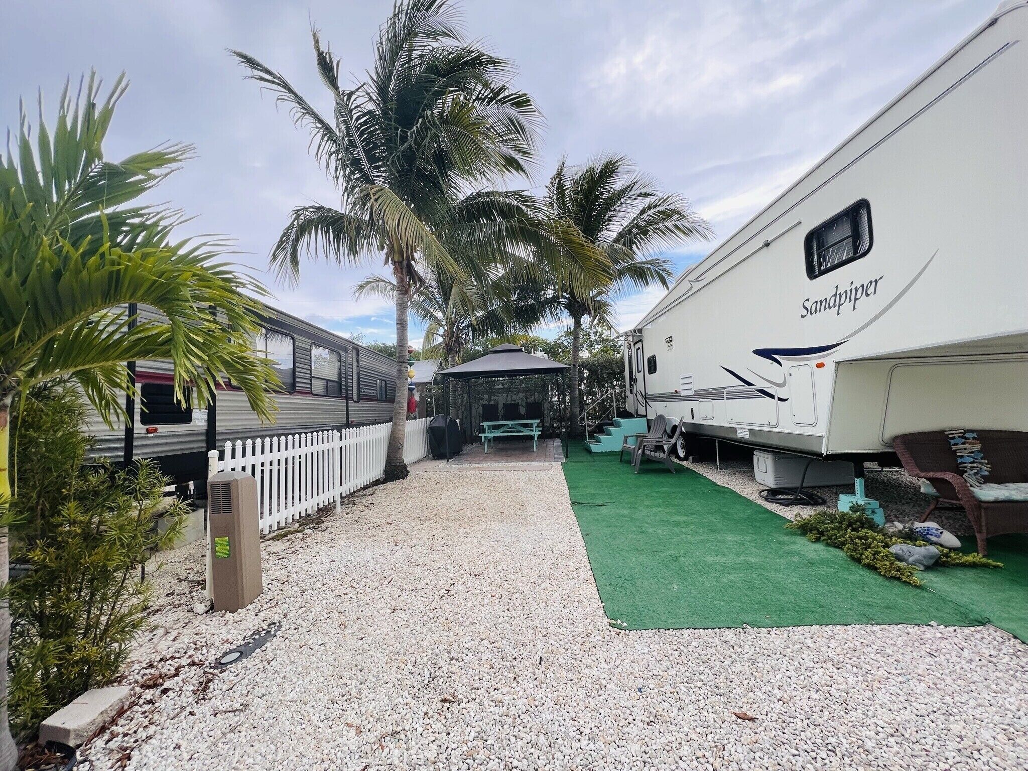 A recreational vehicle (RV) named 'Sandpiper' parked in a campsite with a gravel path, palm trees, and a covered picnic area.