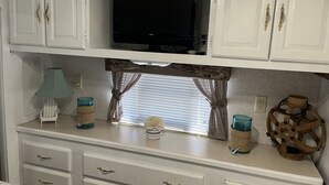Une petite cuisine équipée avec des meubles blancs, un téléviseur, une fenêtre avec des rideaux et une petite table agrémentée d’objets décoratifs.