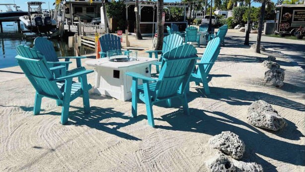 Un área de descanso frente a la playa con sillas Adirondack turquesas y una pequeña mesa, rodeada de arena y palmeras.