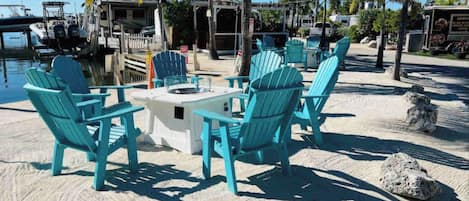 A beachside seating area with turquoise Adirondack chairs and a small table, surrounded by sand and palm trees.
