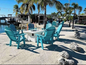 A beachside seating area with turquoise Adirondack chairs and a small table, surrounded by sand and palm trees.