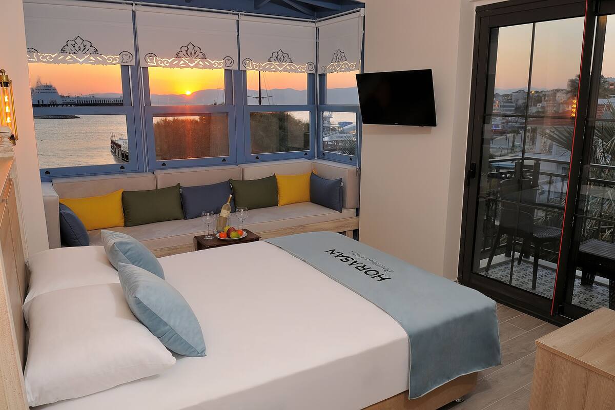 A hotel room with a bed, a sofa, a small table with glasses and fruit, and a balcony with a view of the sunset and cityscape.
