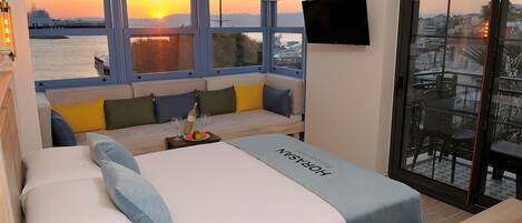 A hotel room with a bed, a sofa, a small table with glasses and fruit, and a balcony with a view of the sunset and cityscape.
