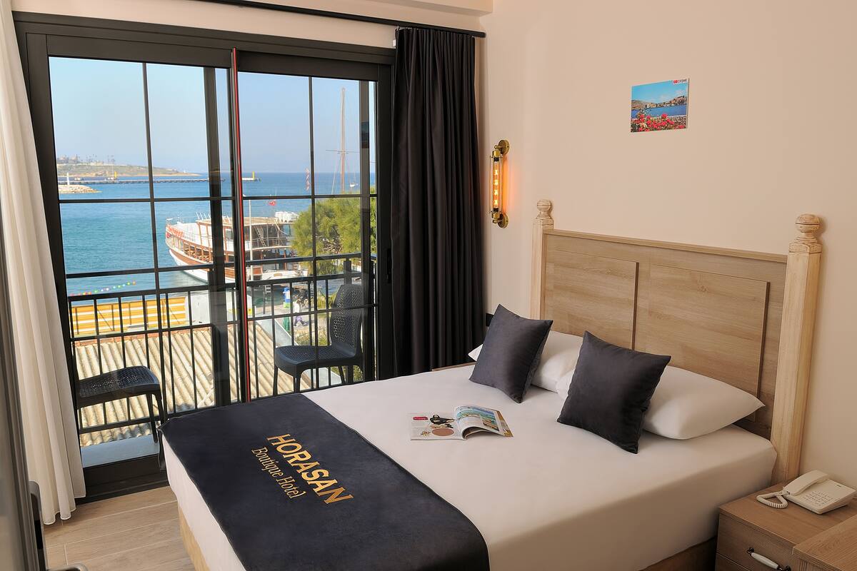 A hotel room with a large window, a bed with a black headboard, and a view of the sea and boats.