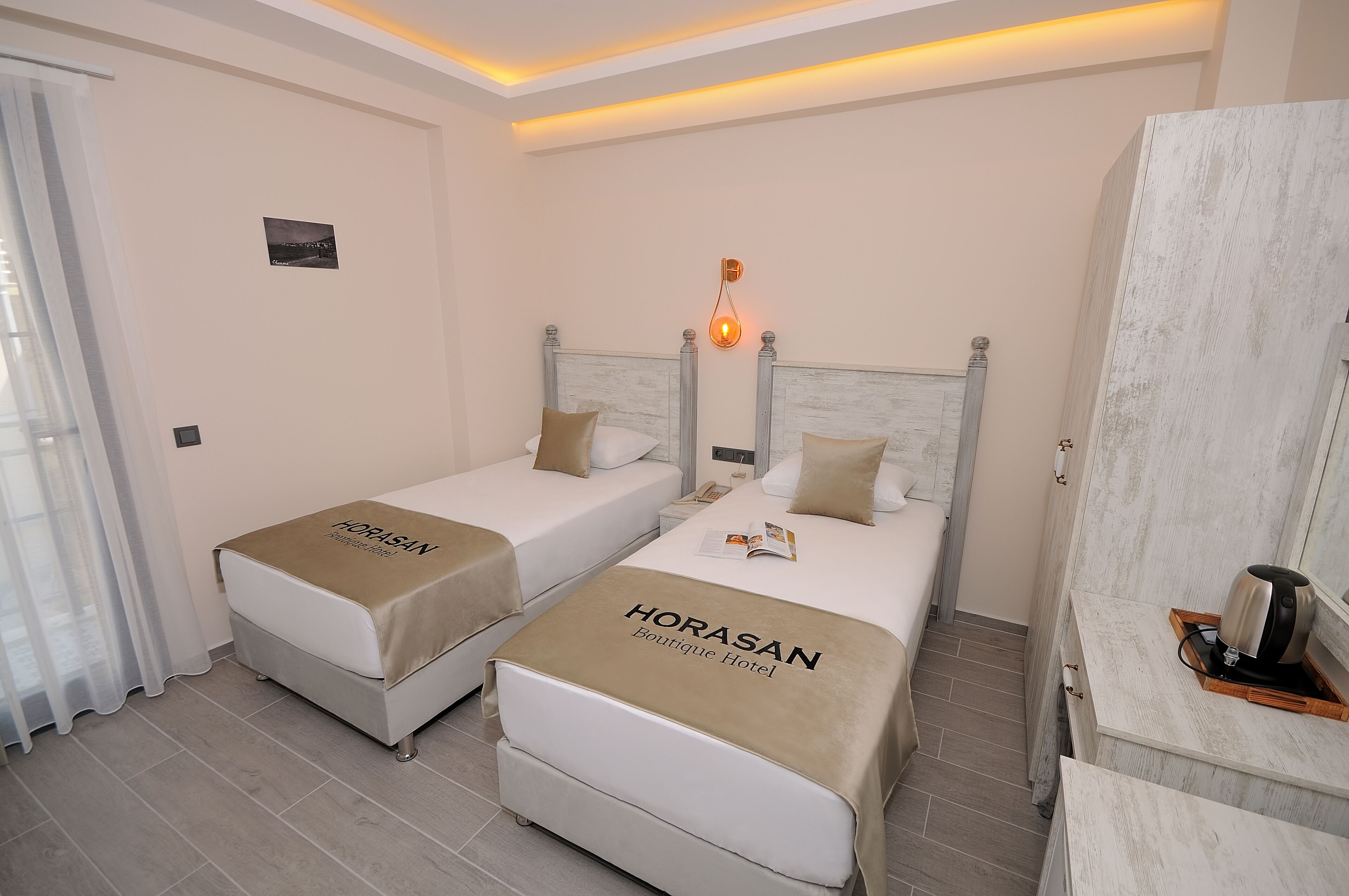 Habitación de hotel con dos camas, cada una con cubrecama del 'HORASAN Boutique Hotel'. Se observa una mesita de noche con lámpara, una pequeña foto en la pared y una ventana con cortinas.