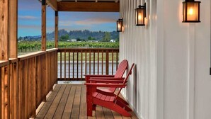 Eine Holzterrasse mit einem roten Adirondack-Sessel, eine weiße Wand mit zwei Wandleuchten und Blick auf einen Weinberg.