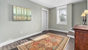 Un couloir avec un tapis à motifs, un tableau accroché au mur, une porte, une fenêtre avec des stores et une lampe posée sur une table.